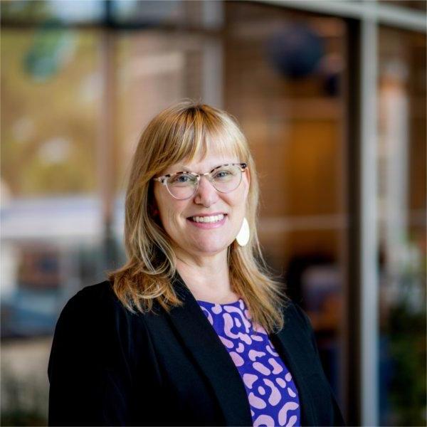 A portrait of Holly Jasinski, a woman with blonde hair and glasses. She smiles and poses in front of the Center for Women and Gender Equity.